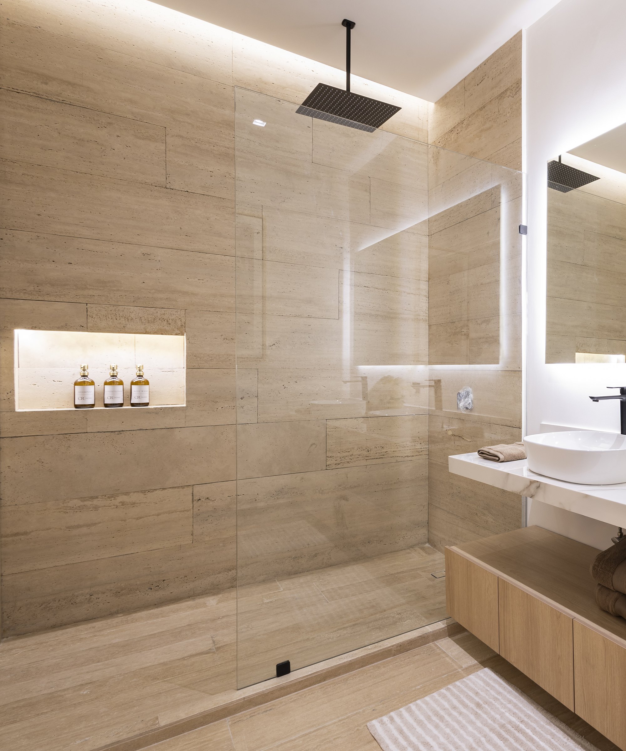 Travertine walk-in shower with a black rain shower head and a lit niche